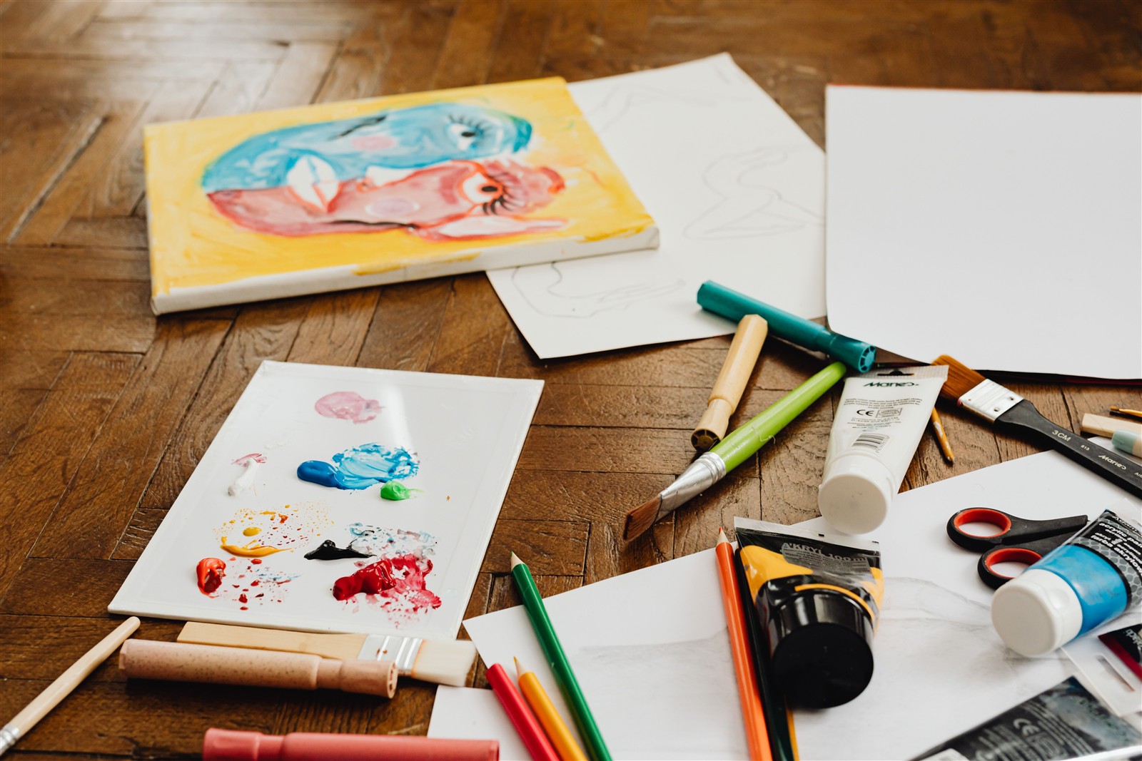 Paint, pencils, brushes, and unfinished artwork spread across a wooden table